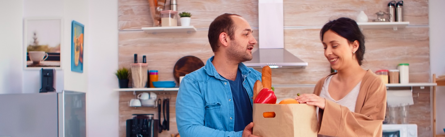 Header: Ein Mann und eine Frau packen gemeinsam einen Lebensmitteleinkauf in einer Küche aus. 