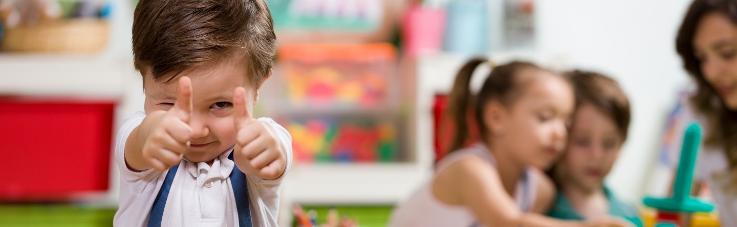 Header: Kindergarten Junge gibt Thumbs Up, im Hintergrund Kinder am Tisch