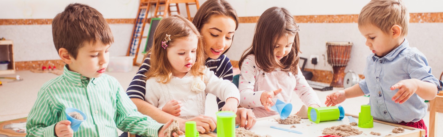 Header: Vier Kinder spielen gemeinsam mit ihrer Betreuerin mit kinetischem Sand. 