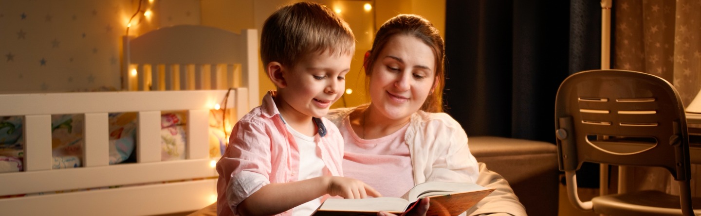 Header: Kinderbetreuerin liest einem Jungen im Kindergartenalter ein Buch vor. Sie sitzen auf dem Boden im Zimmer des Jungen.