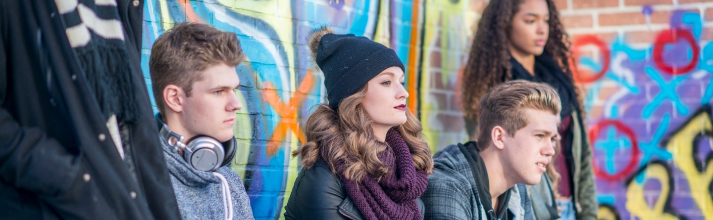 Header: Einige junge Erwachsene sitzen vor einer Wand mit buntem Graffiti.