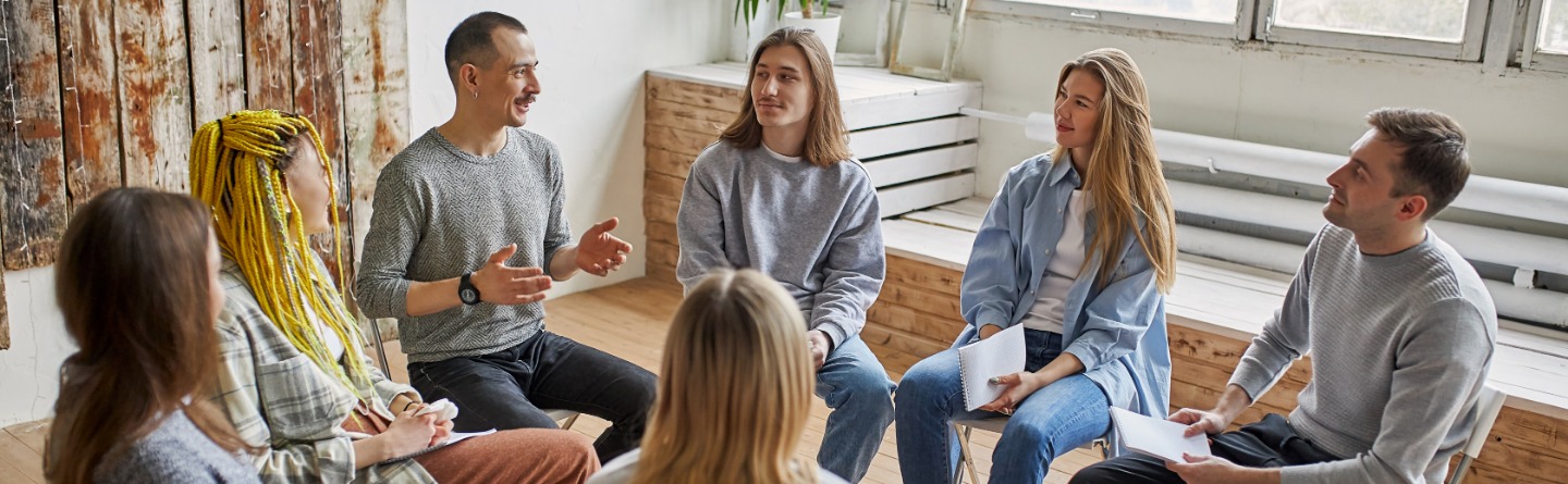 Header: Einige junge Erwachsene sitzen in einem Stuhlkreis und hören einem Mann zu. 