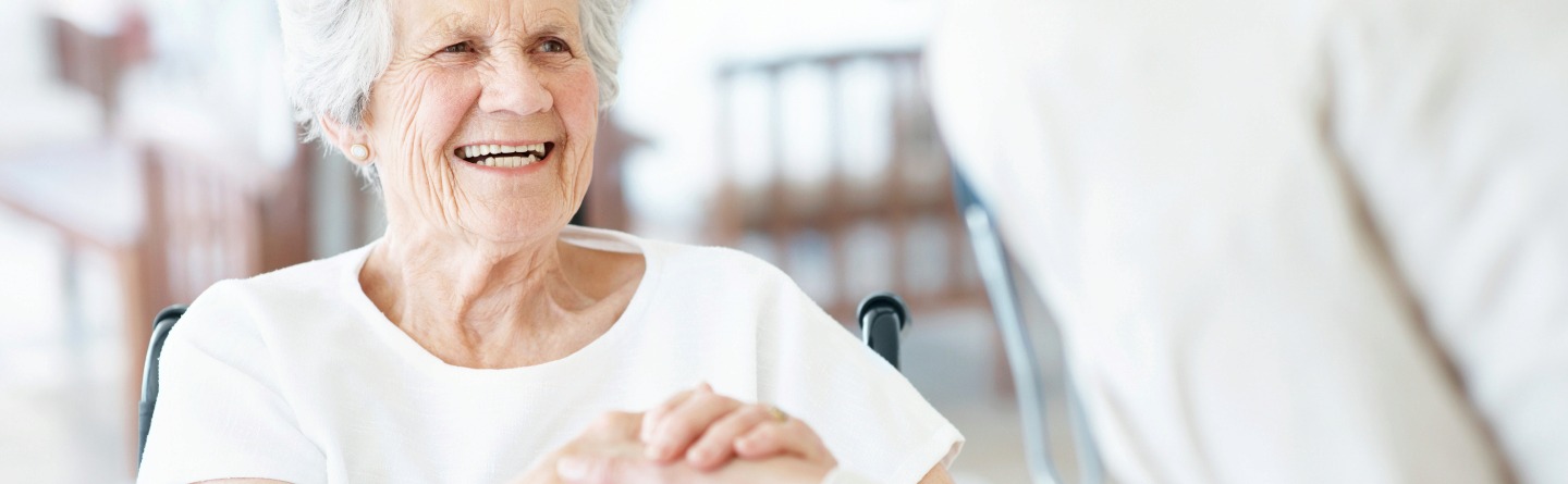 Header: Eine Seniorin schüttelt die Hand ihrer Pflegerin. Dabei lächelt sie glücklich. 