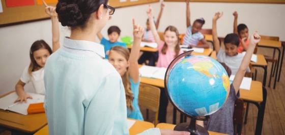 Kachel: Eine Lehrerin zeigt Schülerinnen und Schülern unterschiedlicher Herkunft etwas auf einem Globus. 