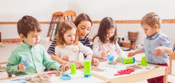 Kachel: Vier Kinder spielen gemeinsam mit ihrer Betreuerin mit kinetischem Sand. 