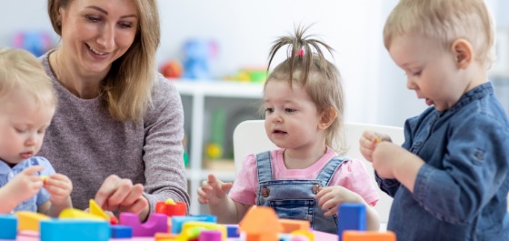Kachel: Drei Kleinkinder spielen gemeinsam mit ihrer Betreuerin mit bunten Bauklötzen