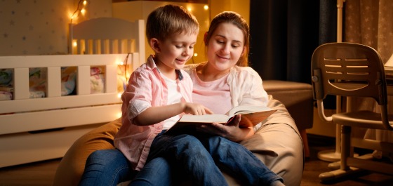 Kachel: Kinderbetreuerin liest einem Jungen im Kindergartenalter ein Buch vor. Sie sitzen auf dem Boden im Zimmer des Jungen.