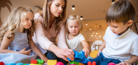 Kachel: Drei Kinder spielen gemeinsam mit ihrer Tagesmutter mit bunten Bausteinen