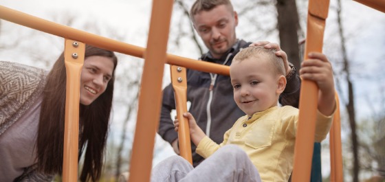 Kachel: Ein Kleinkind spielt gemeinsam mit seinen Eltern auf einem Spielplatz. 
