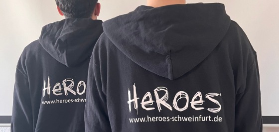 Kachel: zwei Junger Männer sind von hinten zu sehen. Beide tragen einen schwarzen Pulli auf dem groß HEROES steht. 