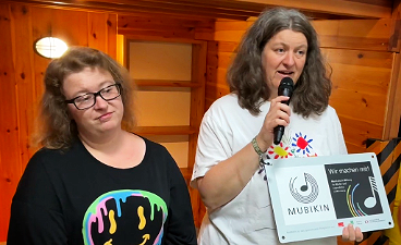 News- Foto: Zwei Frauen stehen nebeneinander, eine hält ein Mikrofon und die MUBIKIN- Förderung in den Händen