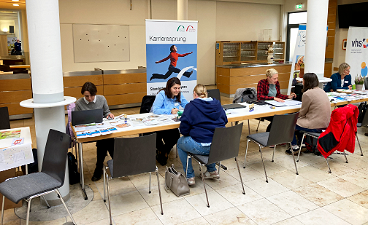 „Woman&Work“: Reges Interesse an Angeboten von bfz und gfi News- Foto: Mehrere Personen sitzen an einer Tischreihe, hinter ihnen ein Rollup