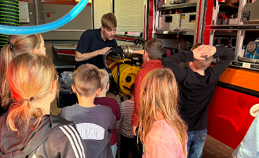 News- Foto: Einige von hinten abgelichtete Kinder beobachten einen Feuerwehrmann, während er ihnen etwas erklärt