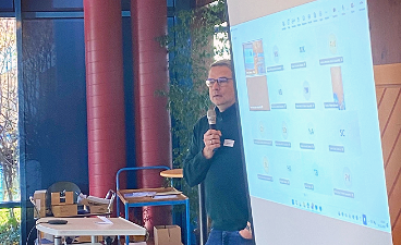 News- Foto: Ein Mann mit Mikrofon steht neben einer Leinwand, auf der eine Präsentation abgespielt wird