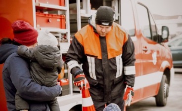 News- Foto: Ein Feuerwehrmann sowie eine Frau, die ein Kleinkind auf dem Arm trägt, stehen vor einem Feuerwehrauto