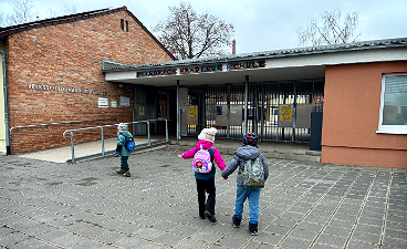 News- Foto: Drei Kinder- zwei davon Händchen haltend- laufen mit ihren Rucksäcken auf das Schulgebäude zu