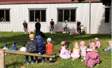 News- Foto: Im Gras sitzende Kinder beobachten die Staffelführer und ihre Hunde