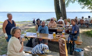 Eine Gruppe sitzt und steht um einen Tisch im Biergarten am Ufer des Starnberger Sees.