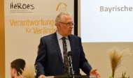 Bayerns Innen- und Integrationsminister Joachim Herrmann hat bei dem Fachtag am gfi-Standort in Schweinfurt dem erfolgreichen Integrationsprojekt zum zehnjährigen Jubiläum gratuliert. Er steht auf einem Podium und hält eine Rede.